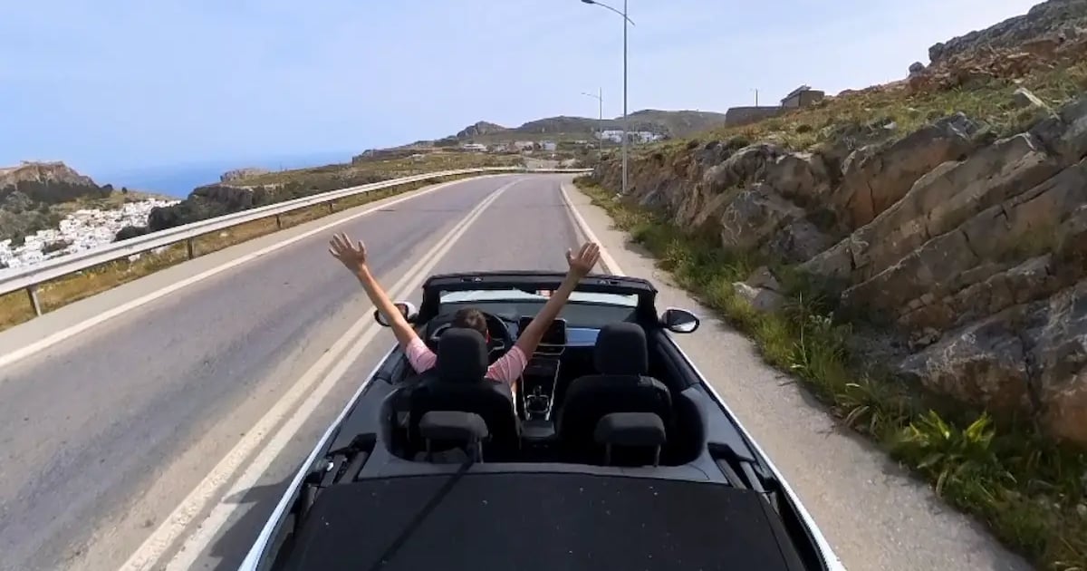 Open-top car on a coastal road in Rhodes, with rocky hills and the sea ahead.