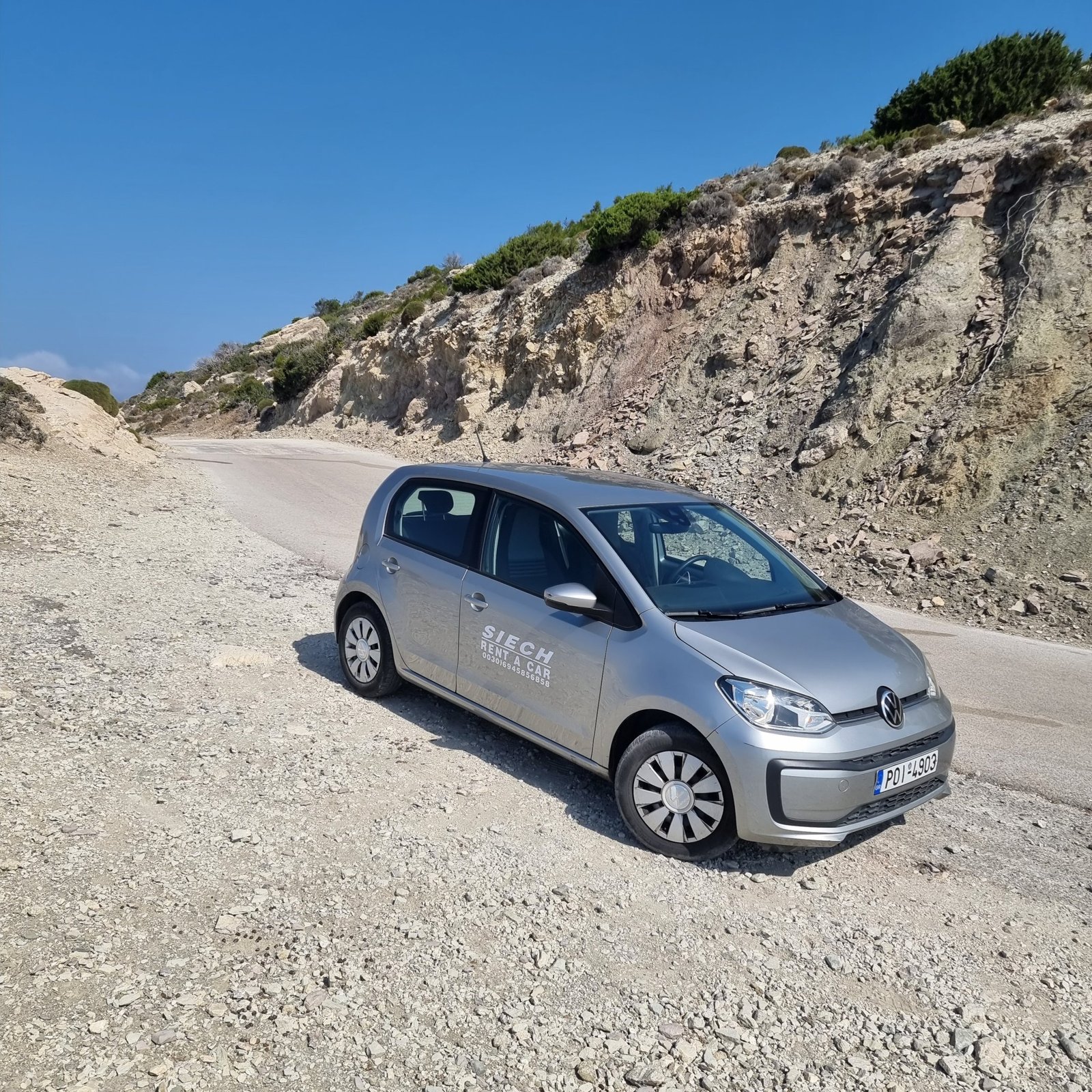 Silver Volkswagen Up from a local Rhodes rental company parked on a gravel roadside below rocky cliffs, photographed by Looneytravels.