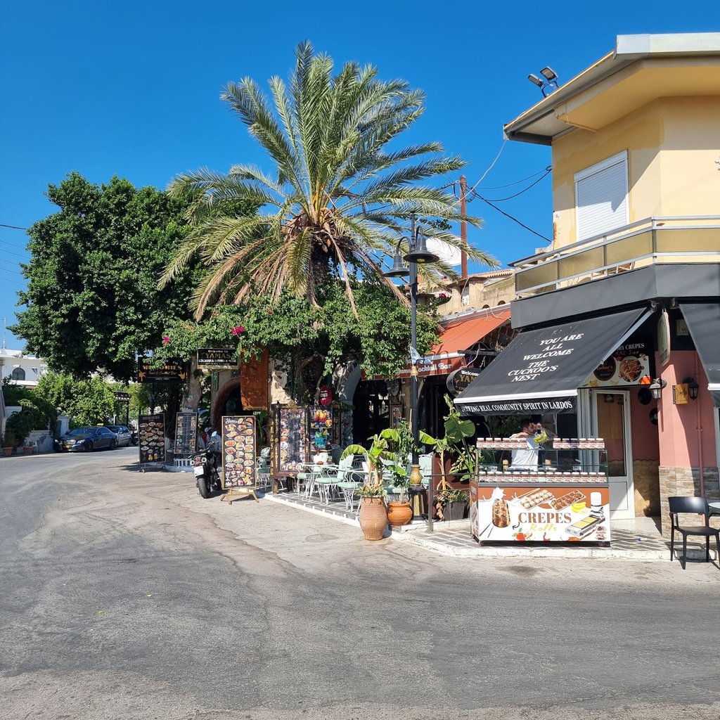Sunny street view of a Rhodes village cafe with outdoor seating, palm trees, and a crepes stand.
